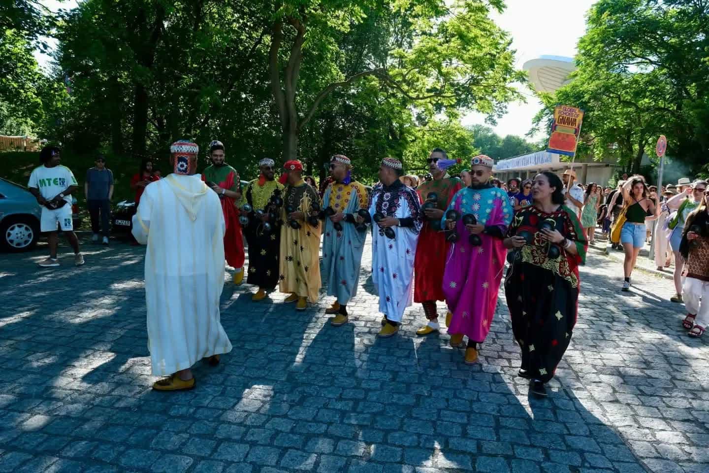The ensemble processes along a cobbled path under trees, a Global Echoes of Gnawa banner raised behind them