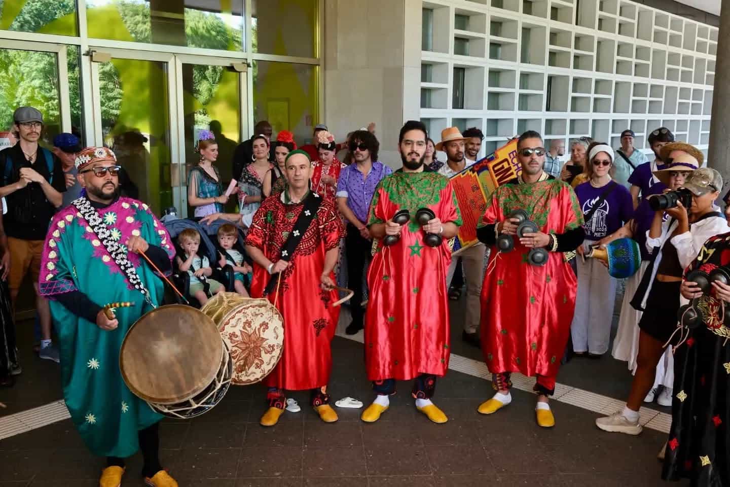 Four Gnawa Berlin musicians in embroidered red and teal robes face the camera with a tbel drum and qraqeb, a crowd behind them