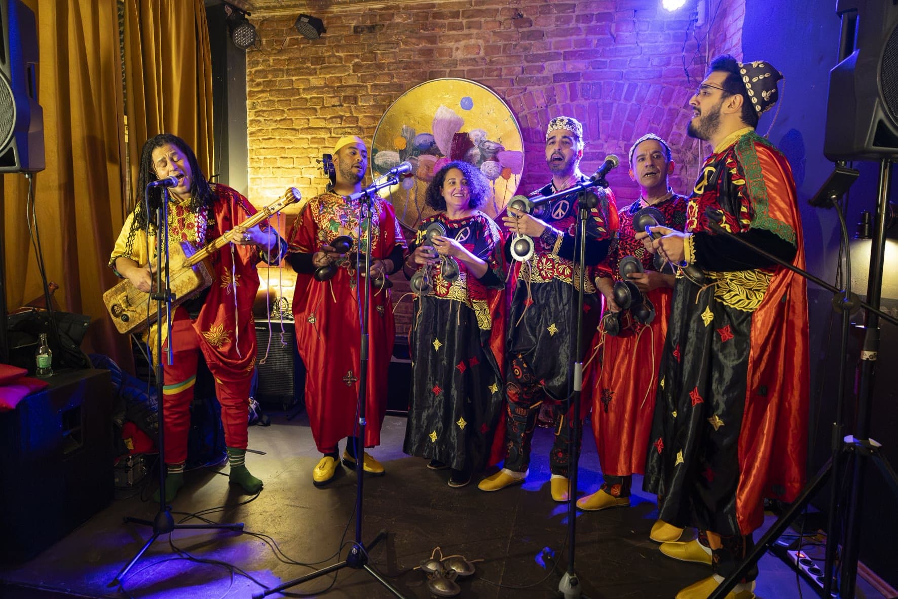 The full ensemble performing on stage against a brick wall and gold curtain under purple light, the guembri player singing at the microphone