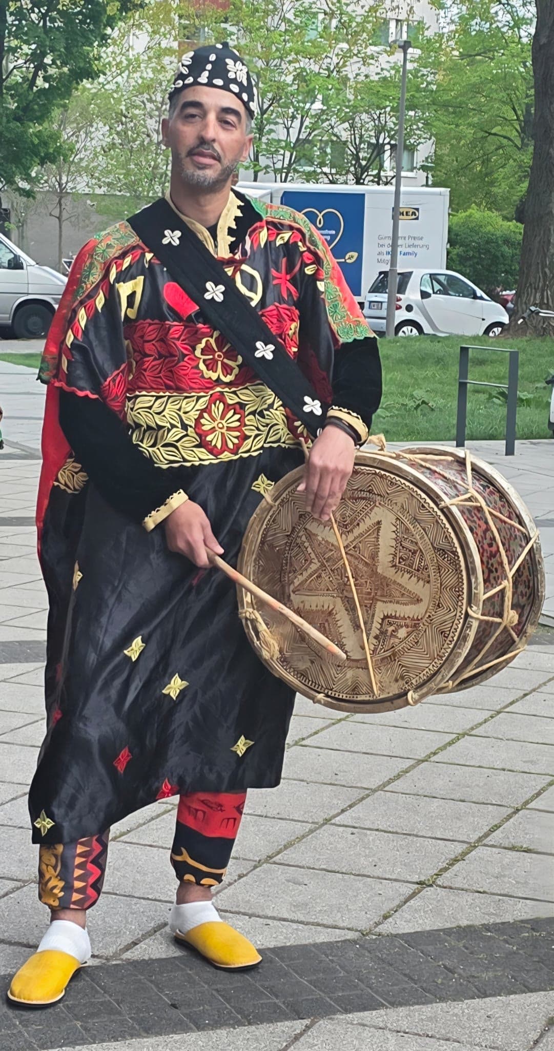 Ein Bandmitglied mit Kaurimuschel-Kappe und schwarz-rot besticktem Gewand hält im Freien eine geschnitzte Tbel-Trommel und einen gebogenen Schlägel